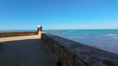 un paseo costero en cádiz con una pequeña caja de vigilancia al final de un muro de piedra, con vistas al océano atlántico bajo un vasto cielo azul