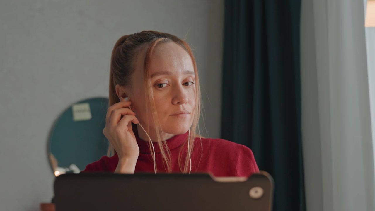 Ung kvinde i rød rullekrave sidder indendørs med åben bærbar computer. Kvinden justerer høretelefoner og kigger ned. En lille potteplante ses kortvarigt i nærheden af hende.