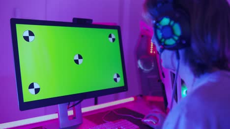 excited gamer playing online video game with a mock up green screen on his powerful personal computer. room and pc have colorful neon led lights. cozy evening at home.