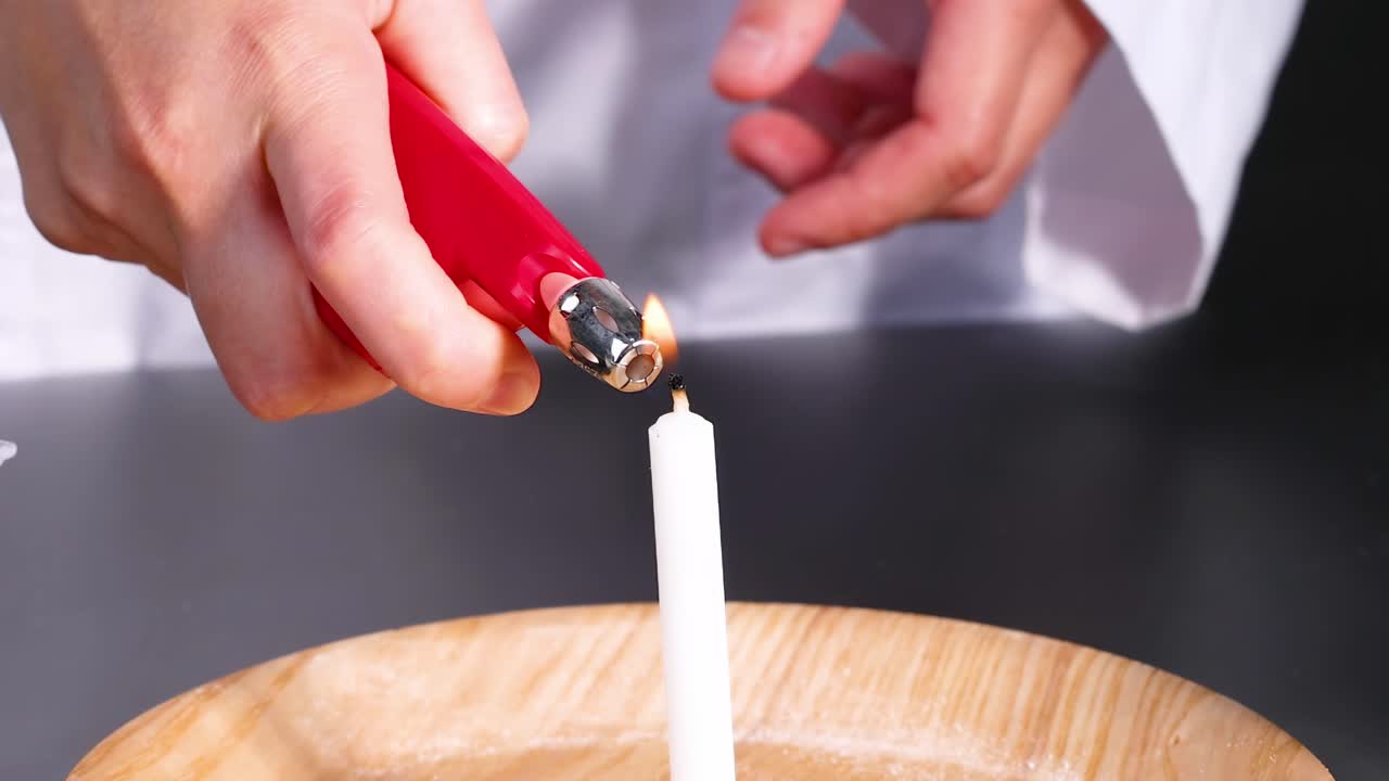A person uses a red lighter to ignite a white candle on a wooden surface.