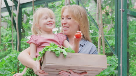 mother and daughter holding box of home grown vegetables in greenhouse