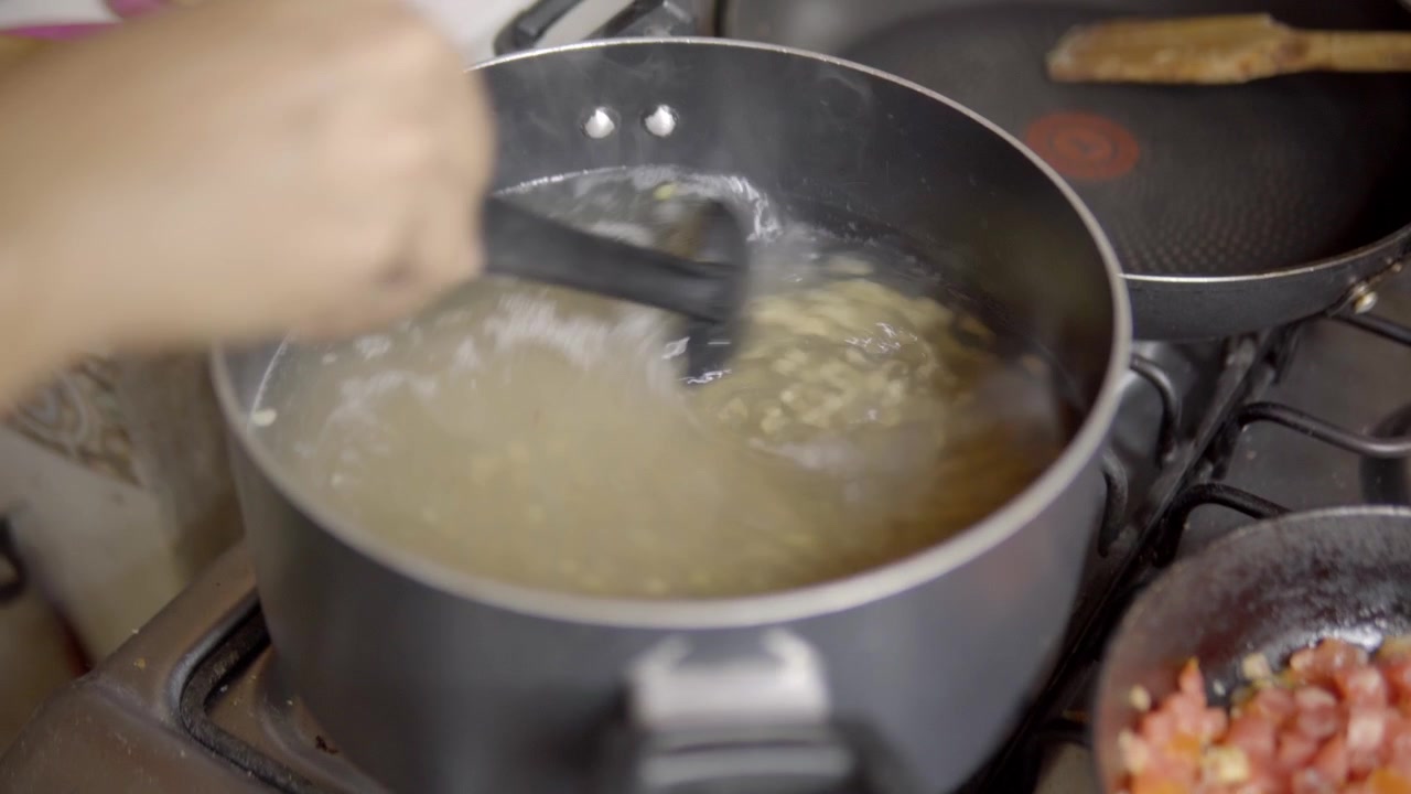 Primo piano di una mano che mescola il brodo che sobbolle in una pentola sul fornello mentre si prepara un pasto fatto in casa, con verdure tritate visibili accanto alla pentola.