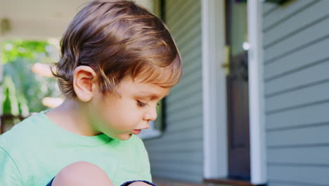 Young-Boy-Sits-On-Porch-Of-House-Playing-With-Toys