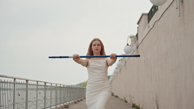 Donna in abito bianco tiene e cammina con una barra blu su un sentiero esterno accanto all'acqua sotto un cielo coperto. Mantiene la stessa postura per tutto il tempo, afferrando saldamente l'oggetto orizzontale