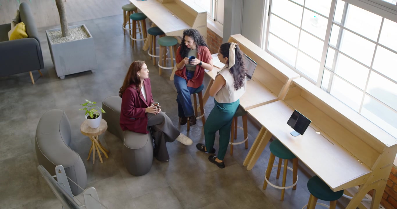 Diverse female coworkers entering coworking lounge from left holding coffee mugs chatting on break