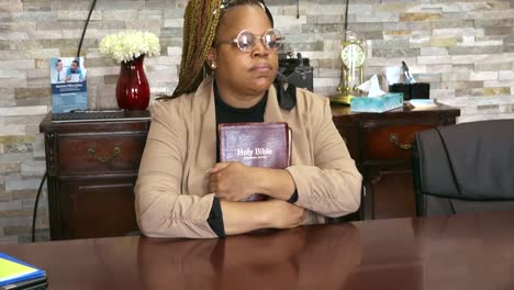 Black-woman-sitting-in-conference-room-mediating-while-holding-bible-and-looking-out-window-on-a-sunny-day