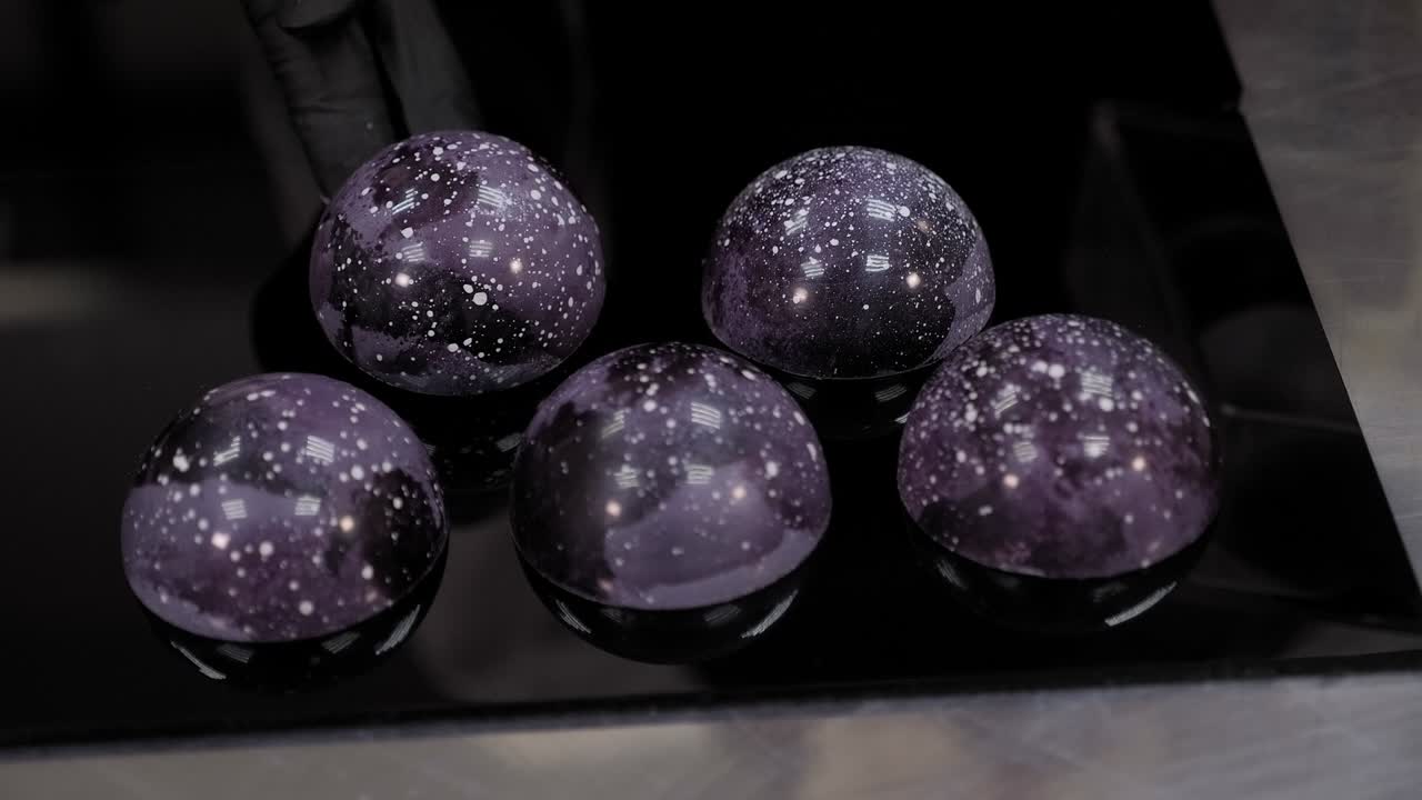 primer plano de un chef de pastelería colocando chocolates de colores en una superficie de espejo negro.