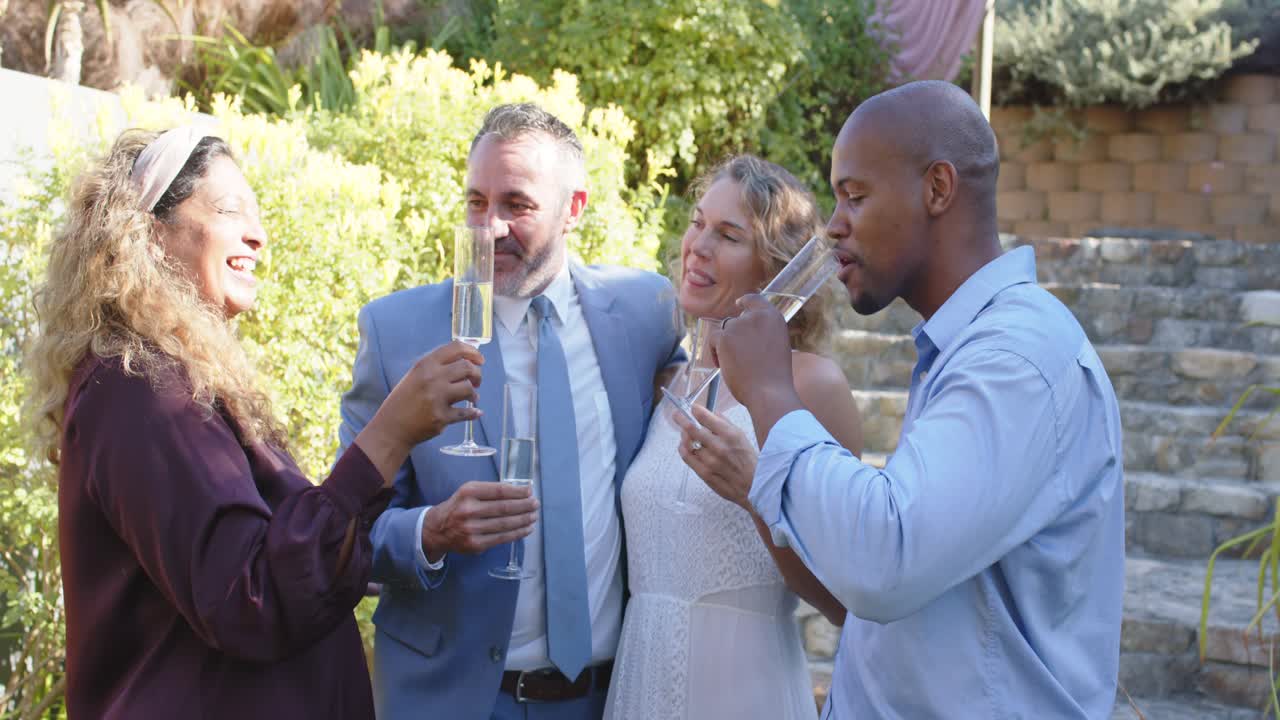 Quartetto eterogeneo che festeggia in abiti formali, alzando flûte di champagne, brindando e sorseggiando in giardino.