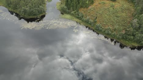 Vista-Aérea-Inclinada-Hacia-Abajo-De-Las-Nubes-Que-Se-Reflejan-En-El-Lago-Todavía-Tranquilo-En-Verano-Noruega-Con-Hierba-Verde-Y-árboles-Que-Se-Revelan-Al-Final-Del-Paisaje