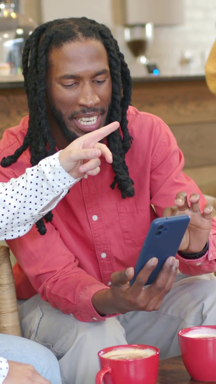 Video verticale: Un braccio a pois indica, mentre un uomo con un top color corallo inclina un telefono blu in un caffè, mostrando delle tazze rosse.