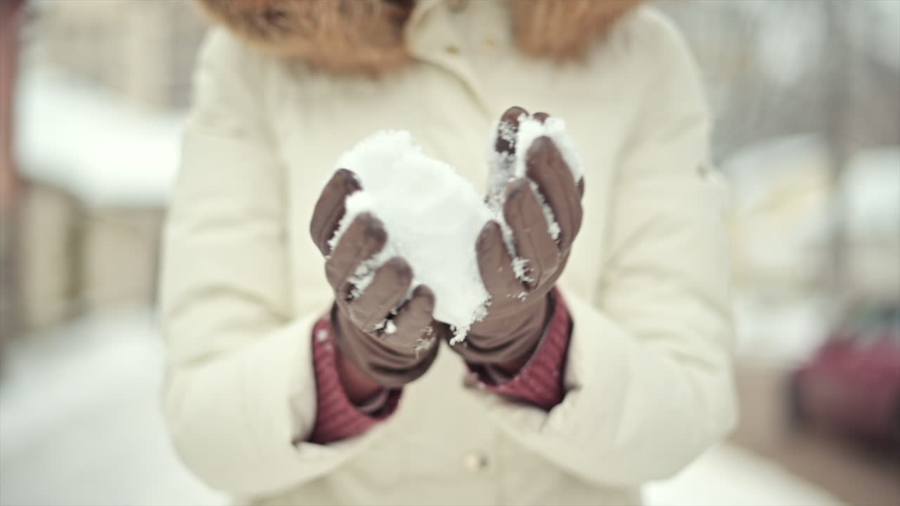 A person with gloved hands scoops up fresh snow, smiling and enjoying the chilly winter day. Snowflakes are gently falling in a quiet neighborhood