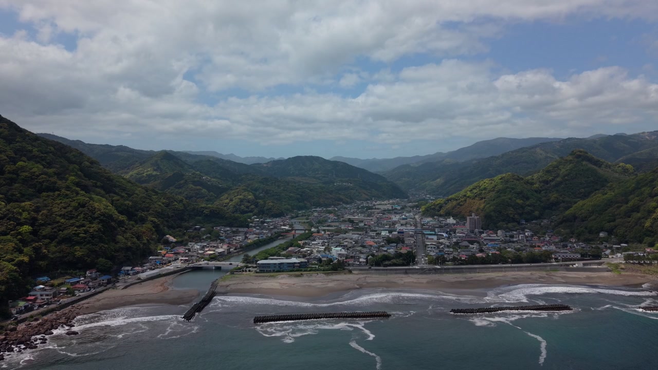Coastal valley town with a river reaching a breakwater lined beach in Kawazu, Izu, Japan, on a partly cloudy day, wide aerial drone shot