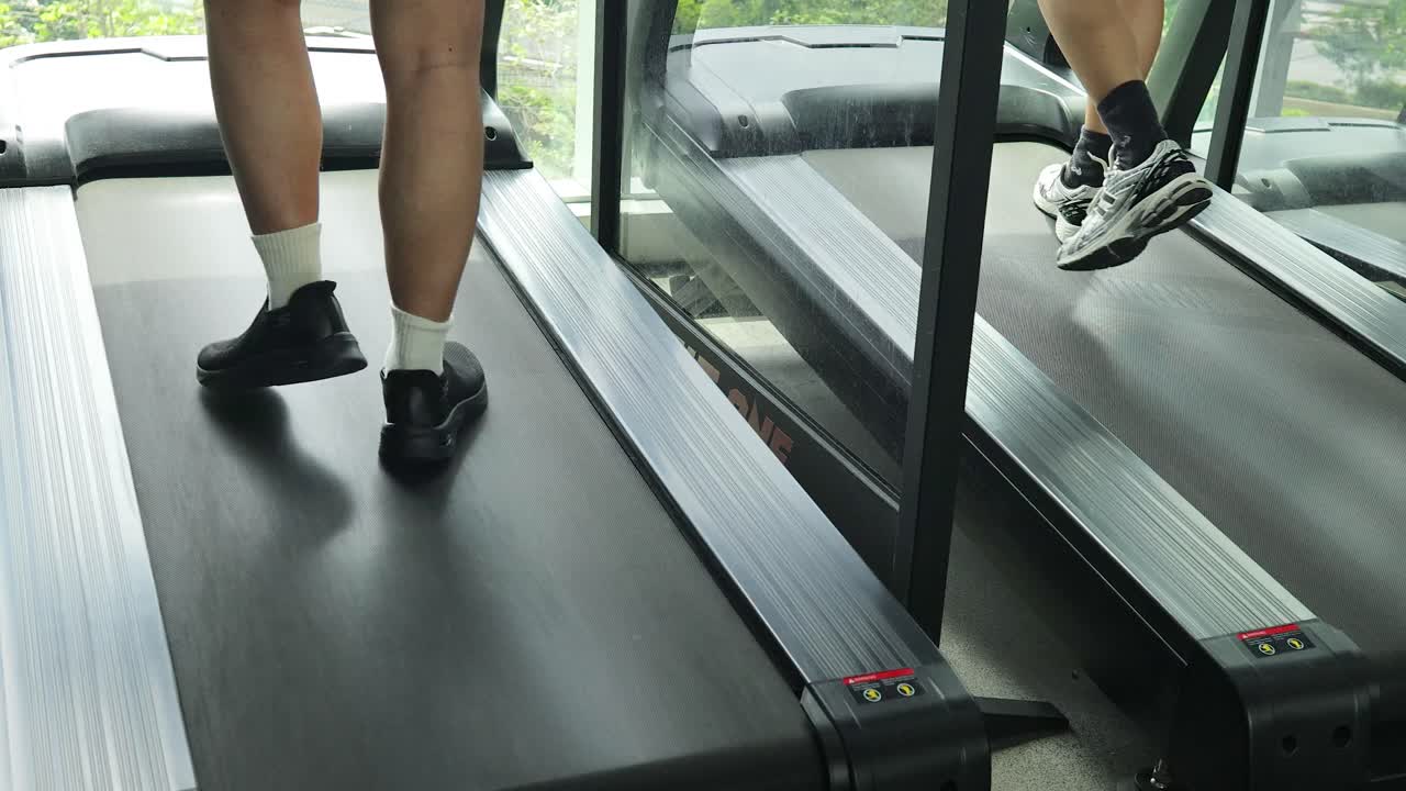 A man in athletic wear walks on a treadmill in a bright, contemporary gym with large windows and natural daylight, captured from a rear angle
