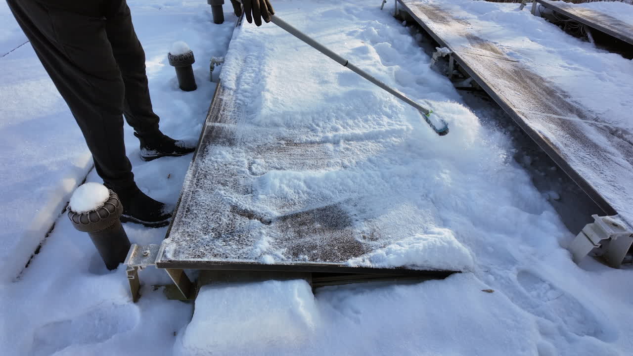 A close-up winter scene showing a person using a broom to remove snow from a rooftop solar panel of a photovoltaic system. The cleared solar module reveals clean glass beneath the thick layer of snow,