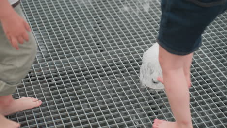 high angle leg view of kids exploring nature closing water fountain jet with feet on metal grate, summer park setting, curious movement, blurred adults and greenery in background