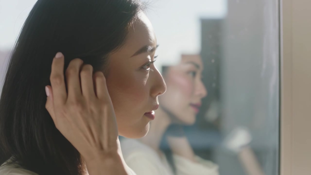 窓際で鏡に映る自分を見ながら、髪を整え、フープピアスやネックレスを直す女性。コピースペースあり。