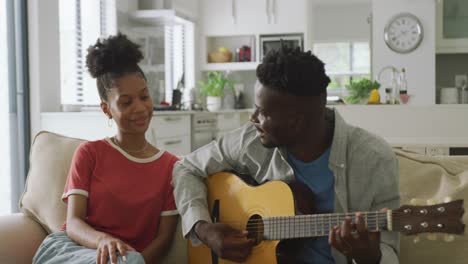 Video-of-happy-african-american-couple-sitting-on-sofa,-singing-and-playing-guitar