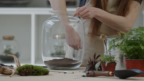 a young woman puts the layer of white sand into a glass jar for creating a tiny live forest ecosystem - close-up