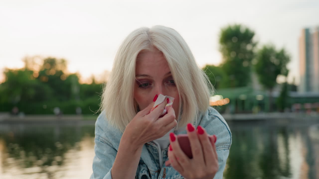 beautiful woman seated on bridge by river park uses compact mirror and white handkerchief to gently wipe bright pink paint from nose after playful painting session during golden hour sunset