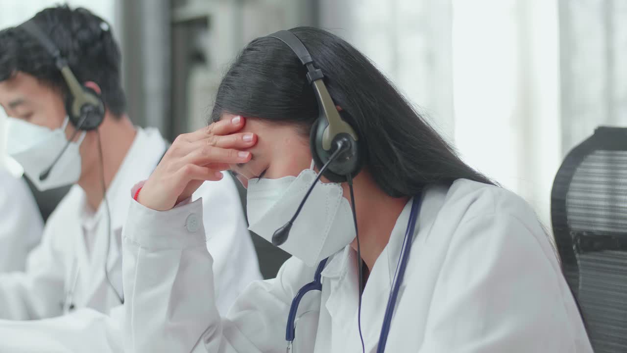 Close Up Of An Asian Woman Doctor Wearing Headsets And Masks Working As Call Center Agent Is Tired Due To Working While Her Colleagues Are Speaking And Typing During A Call At The Office