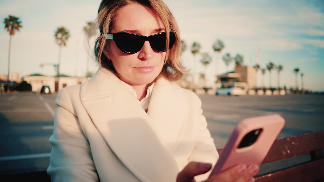 Stylish woman in a white coat looks at her smartphone while sitting on a wooden bench in a sunny parking area lined with palm trees