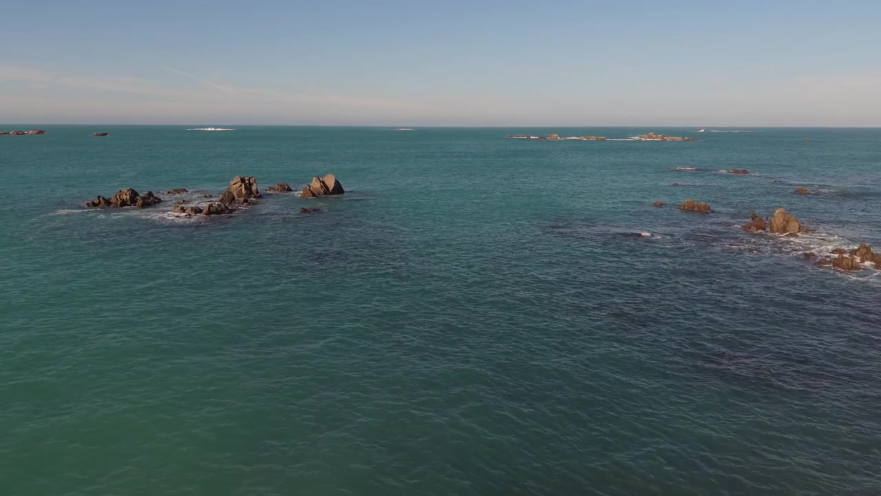 Riprese drone lente e molto basse in avanti verso l'orizzonte sopra acque cristalline e calme intorno a affioramenti rocciosi sotto il sole del tardo pomeriggio, con onde che lambiscono dolcemente le rocce e cielo blu