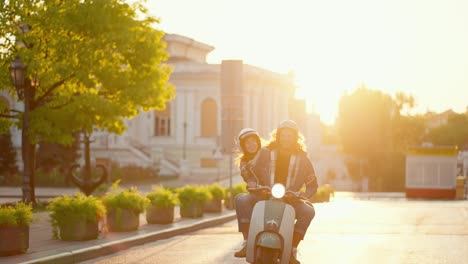 Ein-Glückliches-Paar-Mit-Langen-Lockigen-Haaren,-Einem-Grünen-Motorradhelm-Und-Einer-Jeansjacke-Fährt-Mit-Seiner-Brünetten-Freundin,-Die-Ihm-Hinter-Der-Schulter-Zusieht,-Während-Sie-Auf-Einem-Grünen-Moped-Bei-Einem-Goldenen-Sonnenaufgang-Im-Sommer-Eine-Schöne-Sommerstadtstraße-Entlang-Fährt.