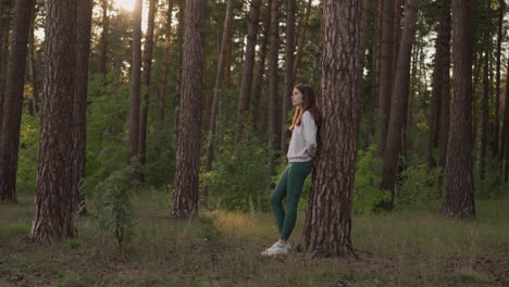 pensive woman rests leaning on tree in forest at sunset. relaxed lady admires beauty of dense trees enjoying sunlight in evening. calmness atmosphere