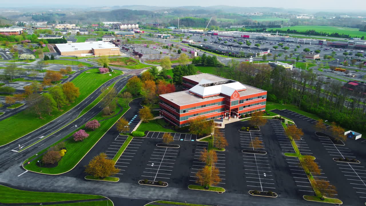 A forward drone pan glides toward a brown brick commercial building in Bucks County, PA, framed by fresh spring greenery, curved roads, and expansive parking lots within a thriving business area.