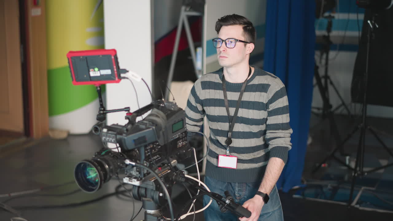 Camera operator focused while adjusting camera with monitor on set. Filming equipment setup in TV studio environment with cables and professional gear