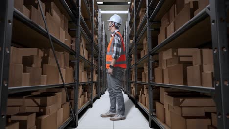 vista lateral completa del cuerpo de un ingeniero masculino asiático con casco de seguridad de pie en el almacén con estantes llenos de mercancías de entrega. mirando a su alrededor, comprobando el stock en los estantes