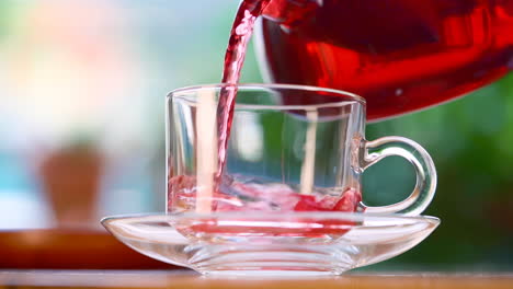 pouring red herbal tea out of transparent tea pot into glass with herbal leaves, lemon and ice cubes with green nature background