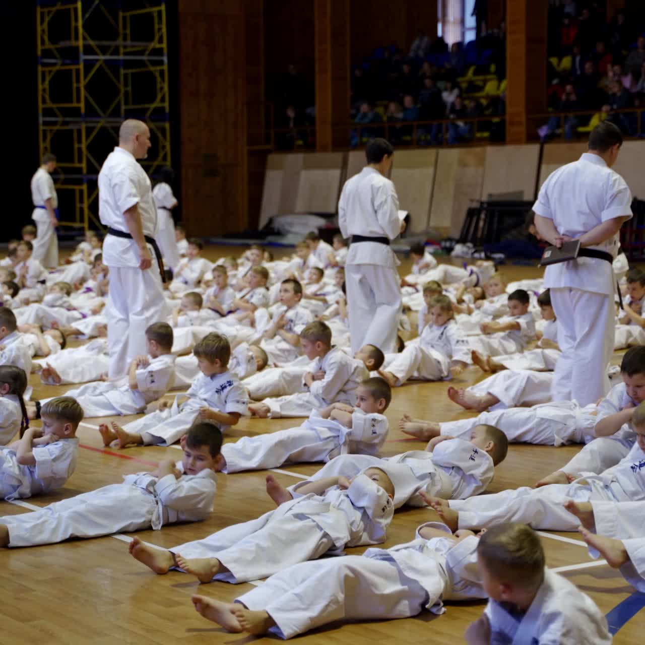 Young karate trainees in kimonos doing crunches in the gym. Coaches control the exercise performance. Audience sitting at backdrop