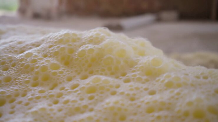 Low near-surface view of dense pale yellow foam filling foreground and midground, with tightly packed bubbles, small cavities, gentle undulations, glossy highlights, and soft creamy texture