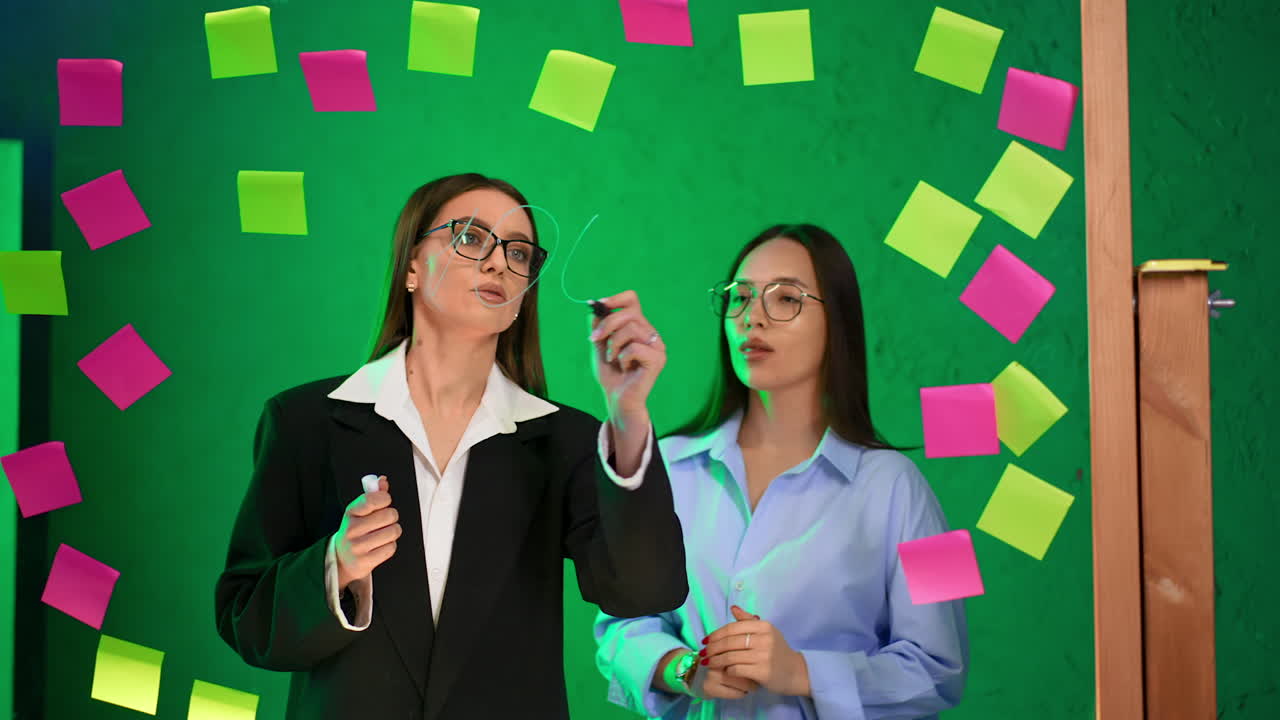 Productive cooperation in the office at glass wall. Brunette ladies discuss strategies, write on the board and consult.