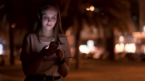 A-young-woman-looks-into-the-smartphone-and-writes-text-messages-on-the-Internet-against-the-backdrop-of-the-night-city.-Girl-businessman-working-on-vacation-remote-work-via-mobile-phone.-Gadget-addiction