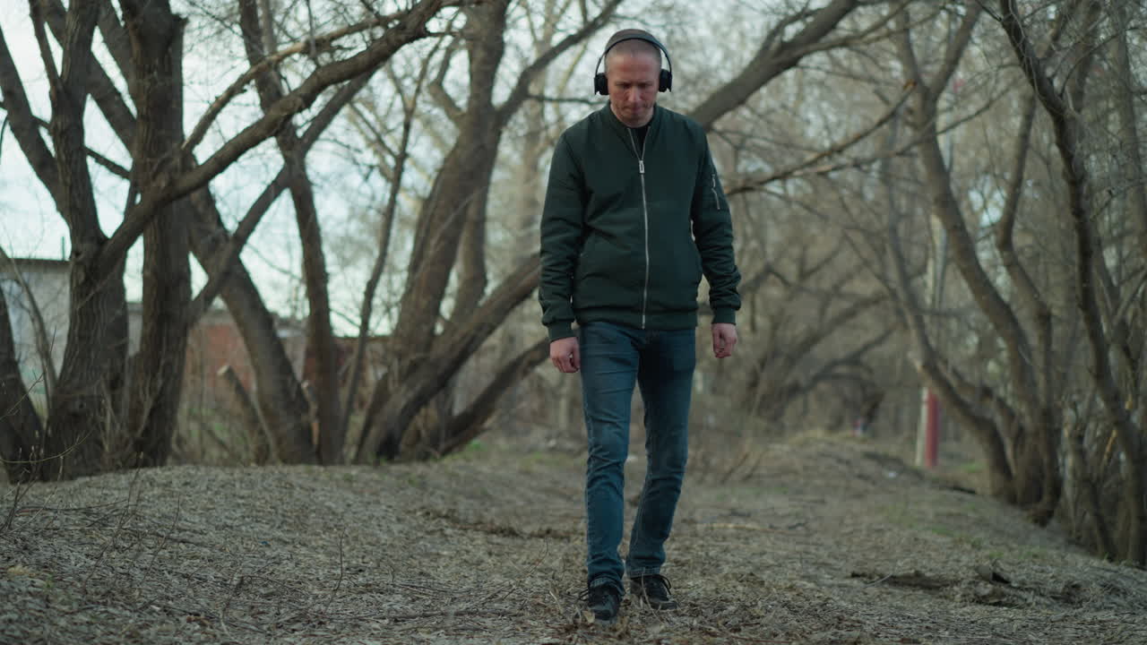 hombre con una chaqueta verde y auriculares caminando solo en un camino sereno del bosque, con el hombre aparentemente perdido en sus pensamientos o disfrutando de su música mientras paseaba por el suelo cubierto de hojas