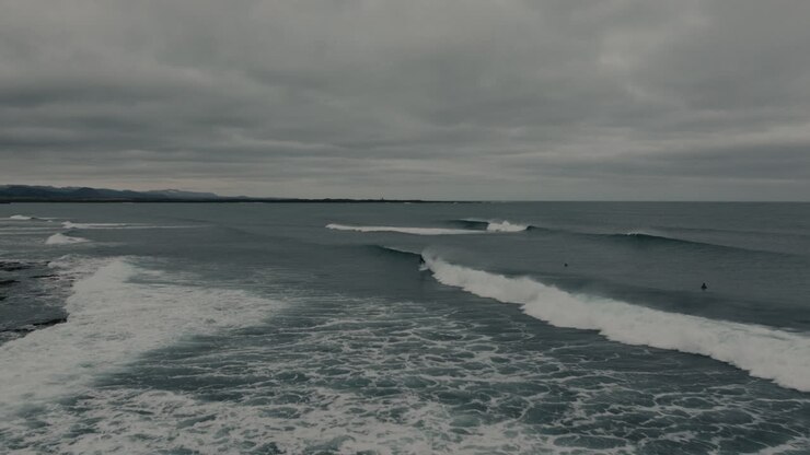 Aerial drone view of a surfer riding a clean ocean wave along the rugged coastline of Iceland under an overcast sky. Cold water surfing, adventure travel and dramatic Nordic landscape concept
