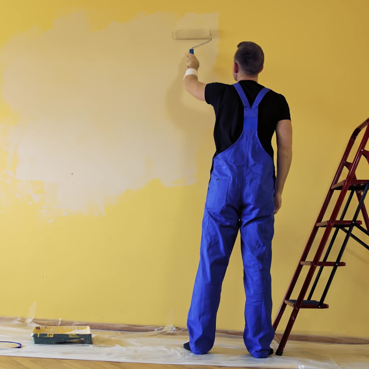 Room decoration with new color. Professional worker in blue overalls painting wall with a paint roller in empty room. Rear view