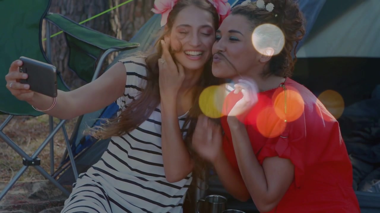 Due donne che si scattano un selfie sociale, la donna a sinistra tiene in mano il telefono, posano con baffi finti, con effetto bokeh fluttuante.