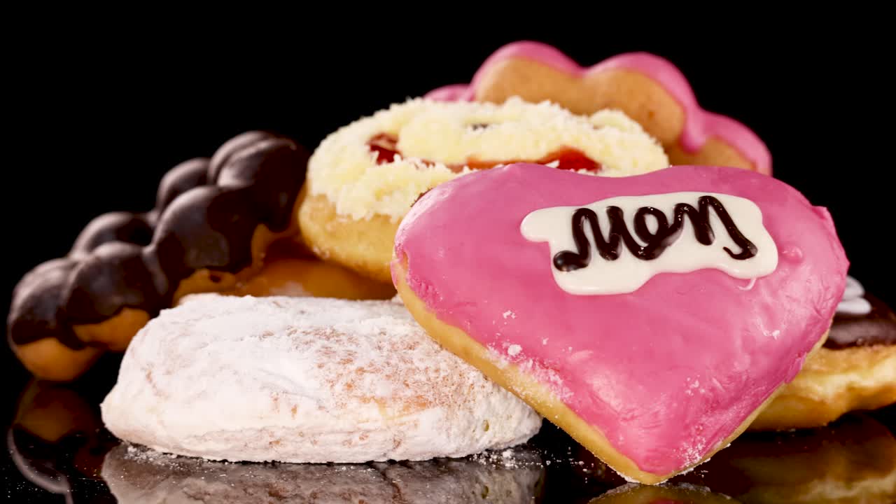 Colorful frosted donuts, heart and ring-shaped, rotate under bright studio lights on glossy black