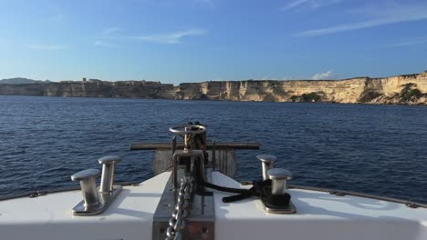 First-person-view-of-bow-of-sailing-boat-moving-forward-navigating-toward-Bonifacio-city-on-top-of-high-cliffs-in-Corsica,-France