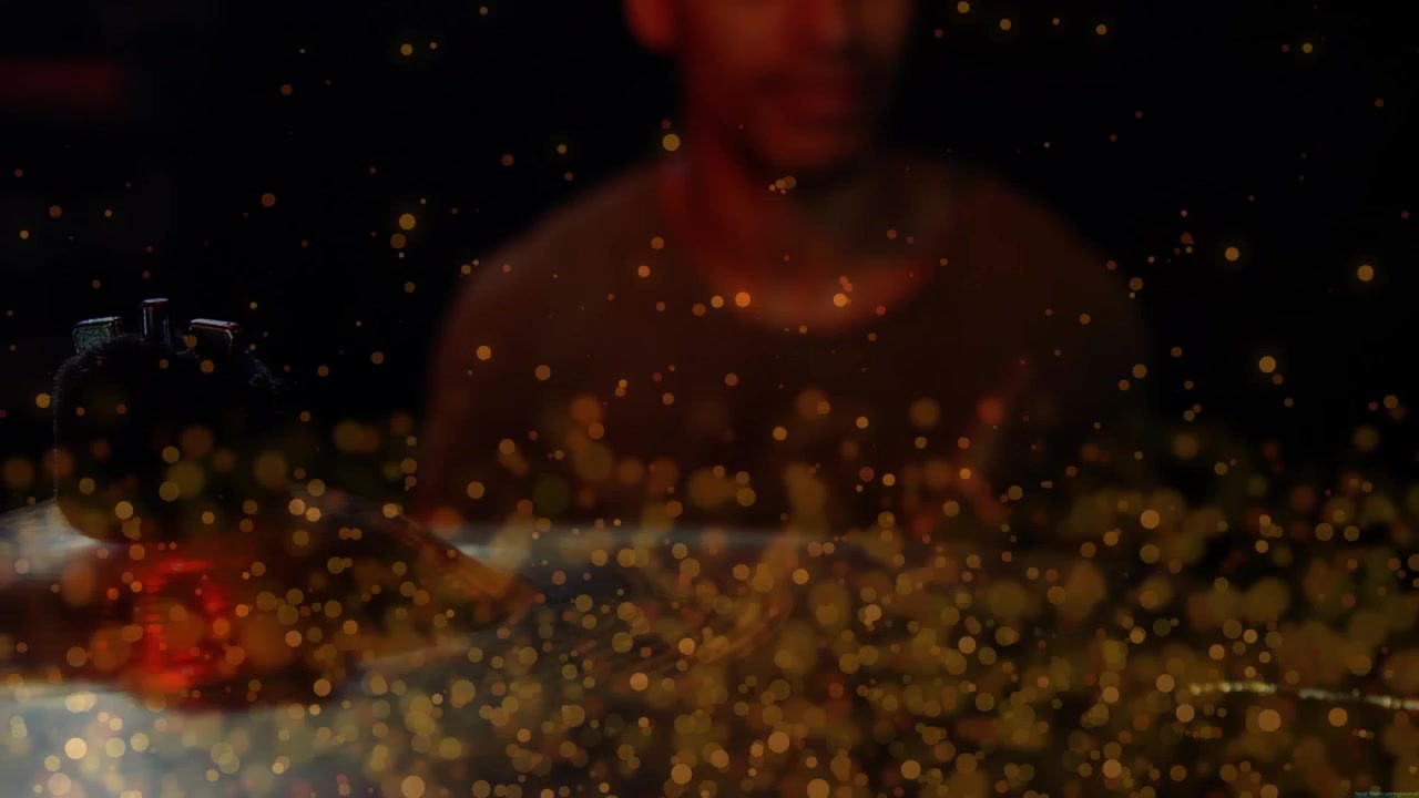Uomo afroamericano seduto in uno studio buio dietro un bokeh dorato in movimento sopra un tavolo, che indossa una maglietta.