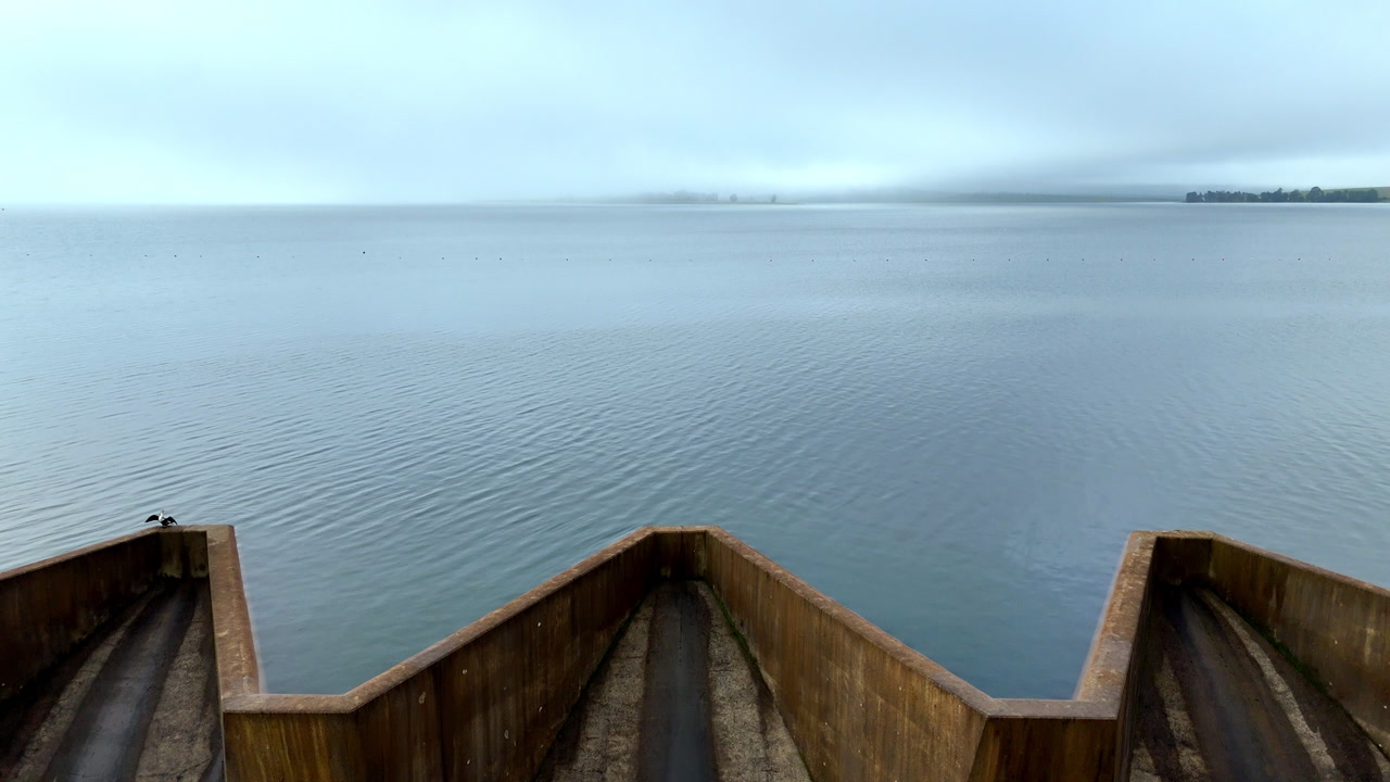Drone rises above concrete dam spillway and calm reservoir