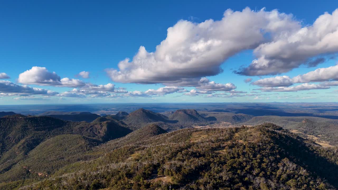 Un dron se desliza suavemente sobre colinas y crestas boscosas en el Parque Nacional Warrumbungle, Australia, bajo nubes dramáticas y luz solar dorada, capturando amplias vistas panorámicas.