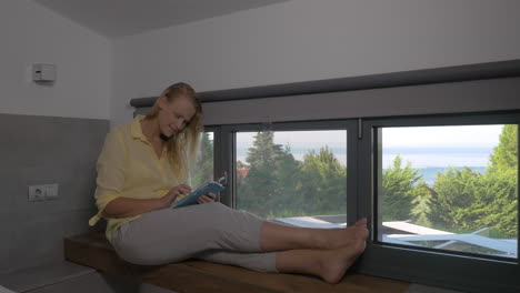 Mujer-Sonriente-Con-Tablet-En-El-Alféizar-De-La-Ventana