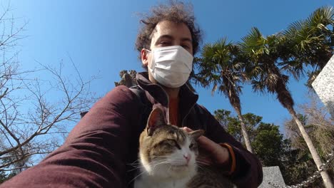 male vlogger holding selfie camera petting cat while wearing facemask