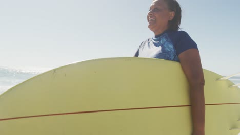 happy senior african american woman walking with surfboard on sunny beach