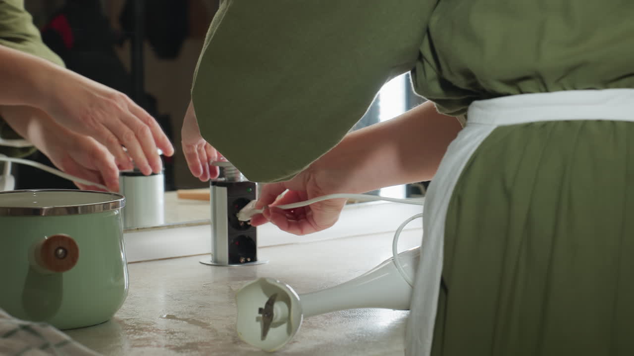 Back view of woman wearing green apron connecting electric hand twister to built-in kitchen socket, preparing to blend ingredients with countertop tools, pastel pot