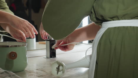 back view of woman wearing green apron connecting electric hand twister to built-in kitchen socket, preparing to blend ingredients with countertop tools, pastel pot